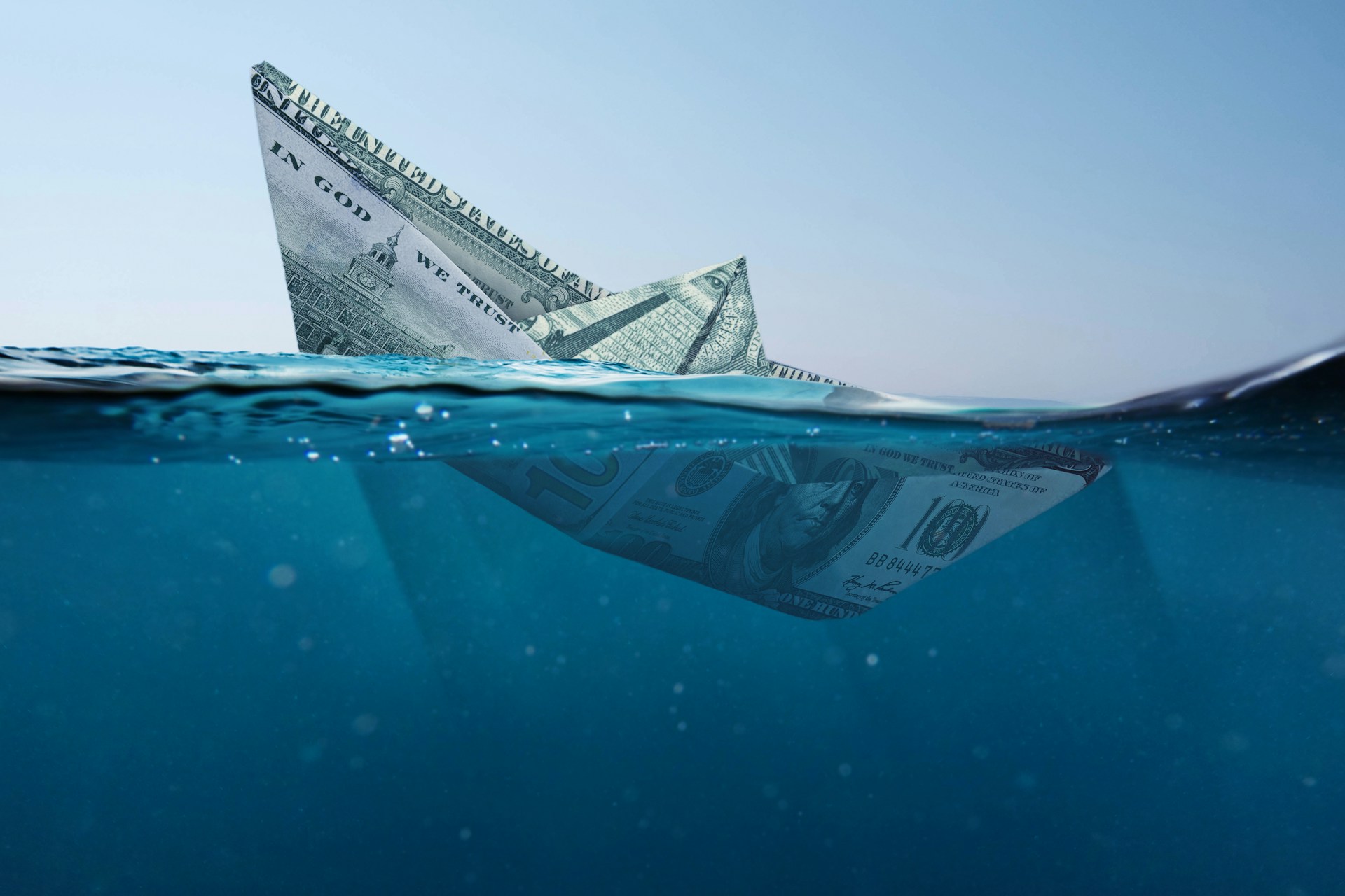 A boat folded from a US dollar bill, half-submerged and sinking beneath the ocean surface